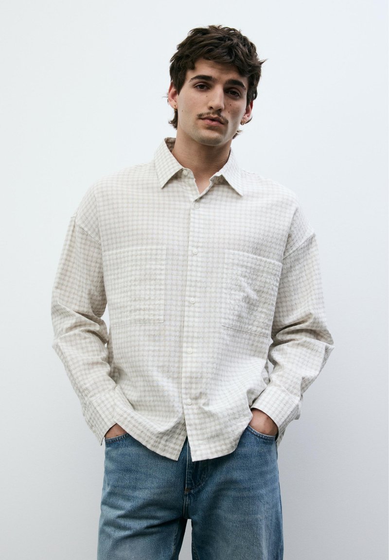 Jeune homme avec une moustache portant une chemise à carreaux gris clair boutonnée et un jean bleu, debout les mains dans les poches devant un fond uni.