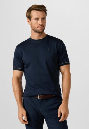Hombre con cabello corto y castaño y barba, que lleva una camisa de manga corta azul marino y pantalones, de pie frente a un fondo claro y liso.