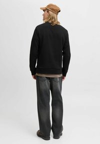 Persona con una gorra marrón, sudadera negra, pantalones vaqueros holgados grises y zapatos marrones de pie, de espaldas, contra un fondo blanco liso.
