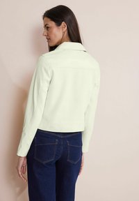 Veste courte vert clair avec un col, des manches longues et un tissu texturé. Associée à un jean en denim bleu foncé avec des poches arrière.