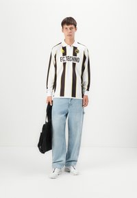 Langermet fotbaltrøye i hvit med svarte og gule vertikale striper, med "F.C.TECHNO"-logo, sammen med lys blå vide jeans og hvite sko.