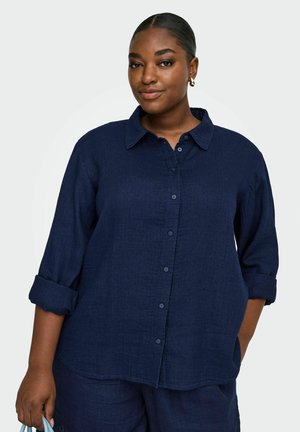 Frau mit mitteldunklem Hautton, die ein marineblaues Hemd mit Knöpfen und passende Hose trägt, ihr Haar zurückgebunden hat und goldene Creolenohrringe trägt.