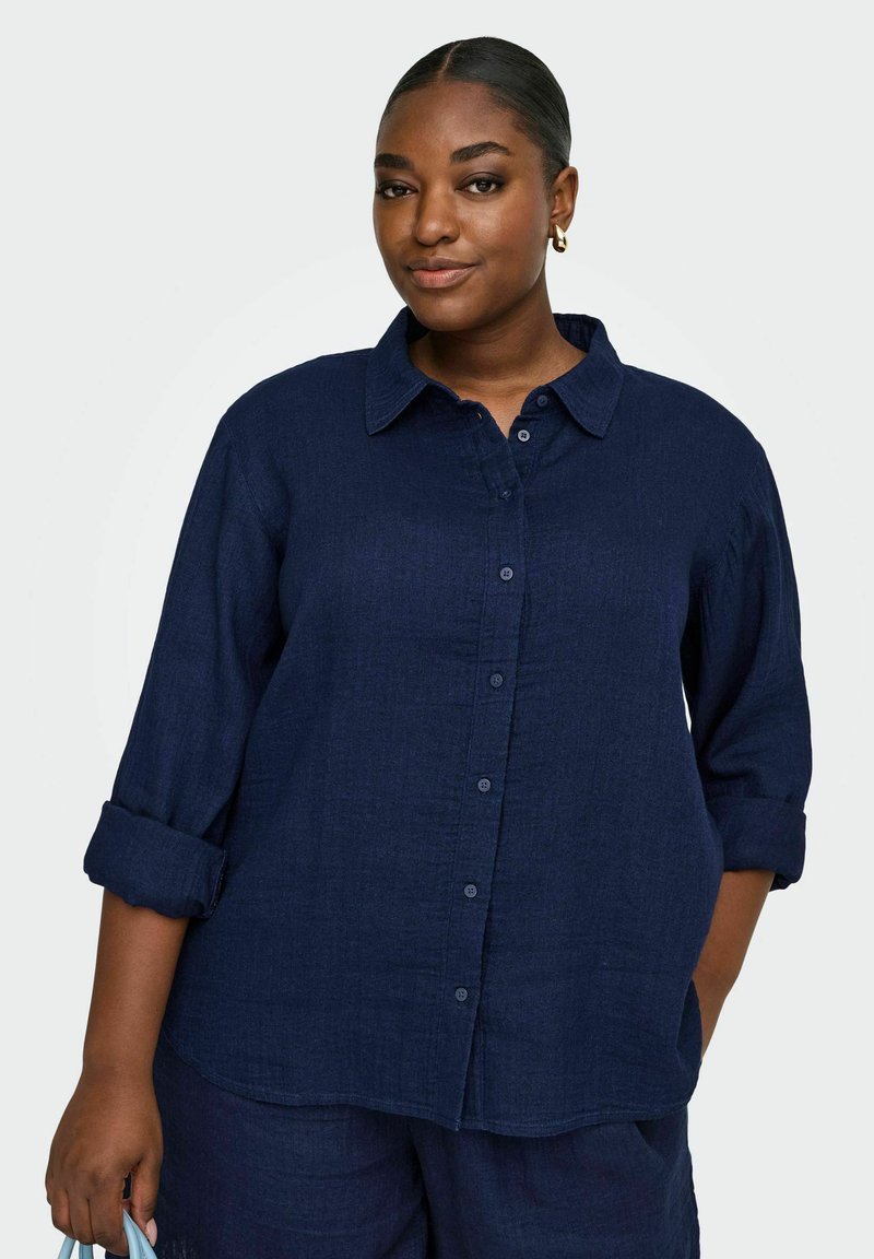 Kvinde med mellem-mørk hudtone iført en mørkeblå skjorte med knapper og matchende bukser, med håret trukket tilbage og guldringe i ørerne.