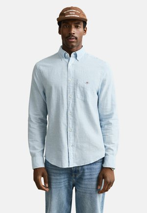 Mann trägt ein hellblaues Hemd mit Knöpfen, blaue Jeans und eine braune Mütze mit der Aufschrift "Saint International Style 1949 American Sportswear."