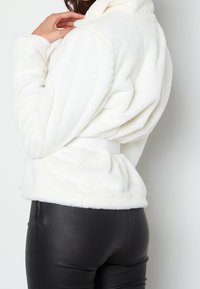 Biała, puszysta kurtka polarowa z wysokim kołnierzem i paskiem w talii, zestawiona z czarnymi, obcisłymi spodniami. Ukazuje widok z tyłu i tekstury.