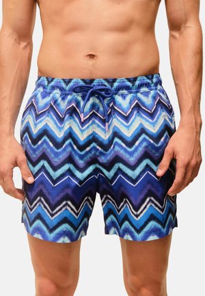 Male torso and upper legs wearing blue and purple zigzag patterned swim shorts with a drawstring waist.