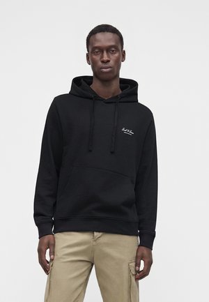 Homme portant un sweat à capuche noir avec poche avant et pantalon cargo beige, debout devant un fond gris uni.