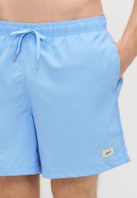 Jasnoniebieskie kąpielówki z elastycznym paskiem w talii, sznurkiem do regulacji, bocznymi kieszeniami i małą naszywką marki na dolnym lewym brzegu.