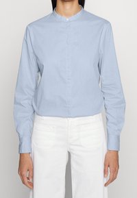 En person som bär en ljusblå långärmad skjorta med mandarinkrage och vita byxor, står mot en neutral bakgrund.