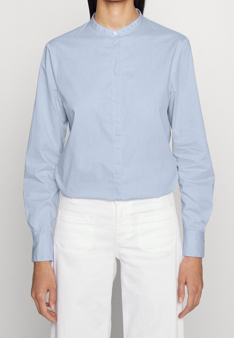 En person som bär en ljusblå långärmad skjorta med mandarinkrage och vita byxor, står mot en neutral bakgrund.