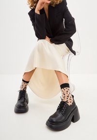 Black platform shoes with a thick sole, paired with sheer leopard print socks. The outfit features a cream-colored satin skirt and a black blazer.