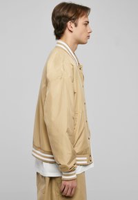 Giacca bomber beige chiaro con polsini e orlo a righe bianche, dotata di chiusura a bottoni a pressione e un fit rilassato.