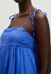Blaues Kleid aus strukturiertem Stoff, mit dünnen Trägern, die an den Schultern gebunden sind, und einem lockeren, gerafften Rock.