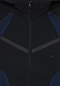 Veste Nike présentant un schéma de couleurs noir et marine, des zips, un tissu texturé, des accents réfléchissants et une capuche au design épuré.