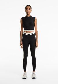 Crni atletski crop top i tajice s bijelim bočnim prugama, izrađeni od glatkog materijala. Imaju uski kroj i visok struk.