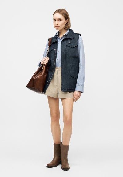 Γυναίκα που φοράει ριγέ πουκάμισο με κουμπιά, σκούρο γιλέκο χωρίς μανίκια, μπεζ σορτς, καφέ μπότες και κρατάει μια μεγάλη καφέ δερμάτινη τσάντα.