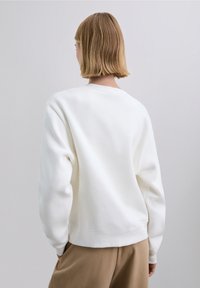 Vit sweatshirt med rund hals, sänkta axlar och ribbade muddar. Mjuk textur med avslappnad passform, kombinerad med beige byxor.