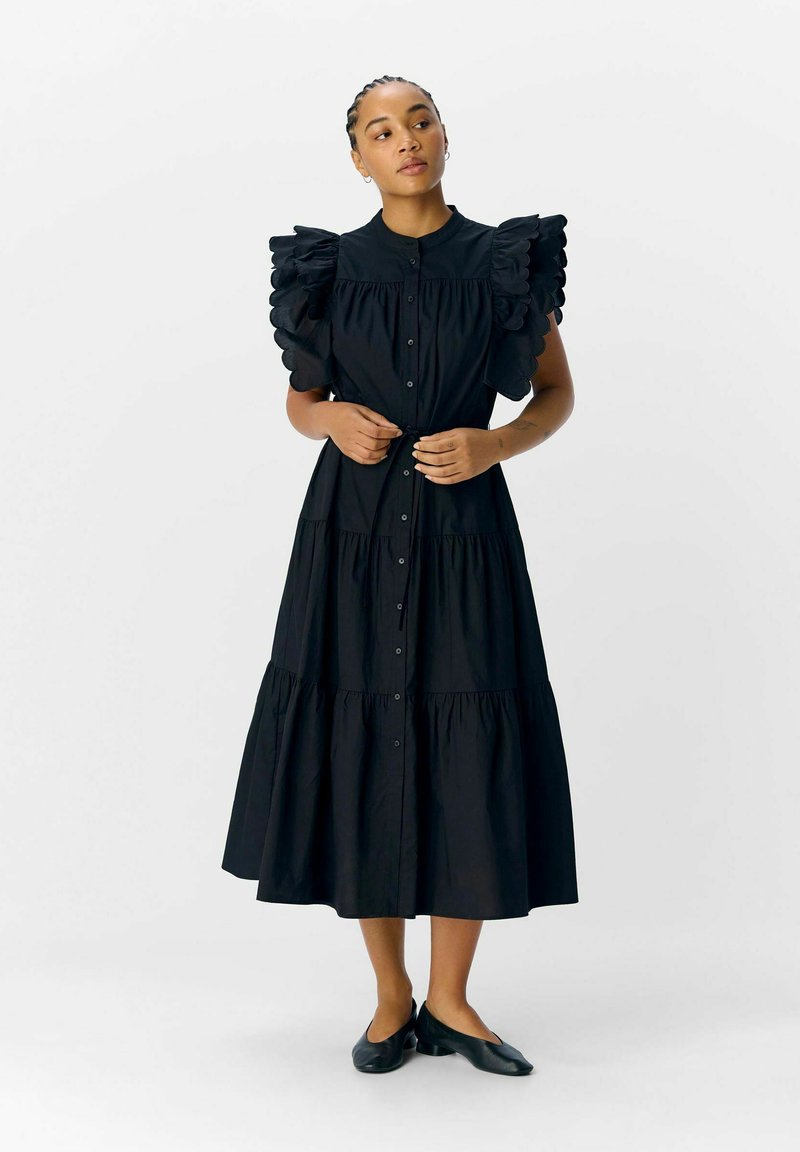 Femme aux cheveux tressés portant une longue robe noire à plusieurs niveaux avec boutons devant et manches courtes à volants, debout les mains sur la taille.