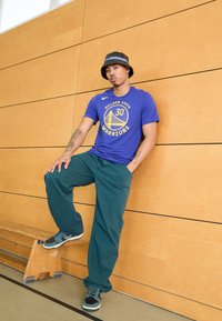 Camiseta azul con el logo de "Golden State Warriors", pantalones verdes y sombrero de cubo negro. Zapatillas negras y verde azulado, banco de madera de fondo.