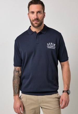 Mężczyzna z brodą i tatuażem na lewej ręce, ubrany w granatową koszulkę polo z napisem „Denim San Francisco” oraz beżowe spodnie, stojący na jasnoszarym tle.