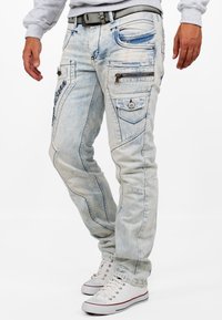 Jeans in denim azzurro chiaro con un aspetto sbiadito, caratterizzati da più tasche, cerniere e dettagli di cucitura. Indossati con sneakers bianche.