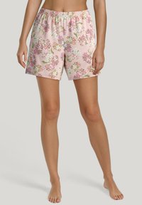 Vrouw in roze bloemenprint shorts met elastische tailleband, staande op blote voeten tegen een effen lichte achtergrond.