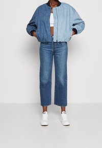 Kurtka bomber z denimu w gradientzie od ciemnoniebieskiego do jasnoniebieskiego, z elastycznymi mankietami i pasem. W połączeniu z spodniami dżinsowymi z wysokim stanem i białymi sneakersami.