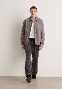 Veste en velours côtelé gris avec col de chemise et logo, associée à un pantalon en velours gris assorti. Modèle portant des chaussures noires. Fond uni.