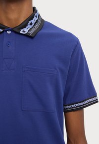 Poloko v žiarivej modrej farbe, s čiernym golierom s modrým reťazovým vzorom a dekoračným lemovaním, vyrobené z textúrovanej tkaniny s náprsníkovou kapsou.