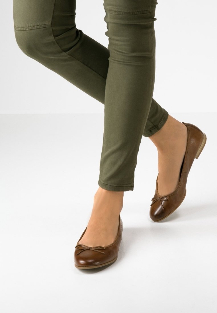 Pieds portant des ballerines en cuir marron avec de petits nœuds, assorties à un pantalon ajusté vert olive sur fond blanc.