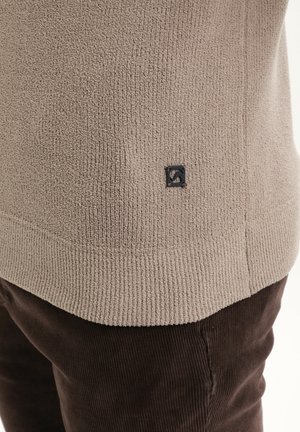 Šviesiai rudas tekstūruotas megztinis su raštuotu kraštu ir maža juoda logotipo etikete šone, derinamas su tamsiai rudomis veliūrinėmis kelnėmis.