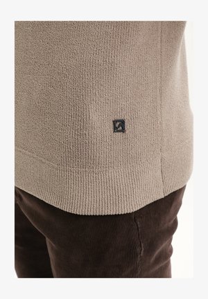Light brown textured sweater with a ribbed hem and a small black logo tag on the side, paired with dark brown corduroy pants.