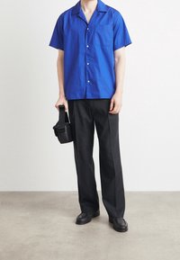 Niebieska koszula z krótkim rękawem i kieszenią, zestawiona z czarnymi spodniami. Czarna torba na ramię i czarne mokasyny dopełniają strój.