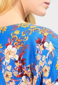 Blusa azul de flores con una textura suave, que presenta flores brillantes en amarillos y rojos, y patrones intrincados de follaje verde a lo largo del escote.