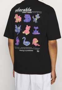 Mustat t-paita, jossa on takana eläväinen graafinen kuva sarjakuvamaista eläimistä eri väreissä. Teksti kuuluu: "suloiset ihanat asiat, kaikki mitä minulla on."