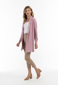 Lang, åben-front cardigan i lys rosa med et struktureret mønster. Kombineret med beige tætsiddende bukser og beige højhælede sko.