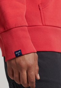 Felpa rossa con polsini a coste e una piccola etichetta blu con il testo "Superdry". La texture del tessuto appare morbida e liscia.
