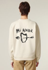 Felpa color crema con testo grafico nero "Mi Amor" e design di un cuore con una freccia sul retro. Tessuto morbido con polsini a costine.
