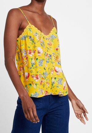 Kobieta ubrana w żółty, kwiecisty top na cienkich ramiączkach z koronkową lamówką, zestawiony z ciemnoniebieskimi dżinsami na jednolitym tle.