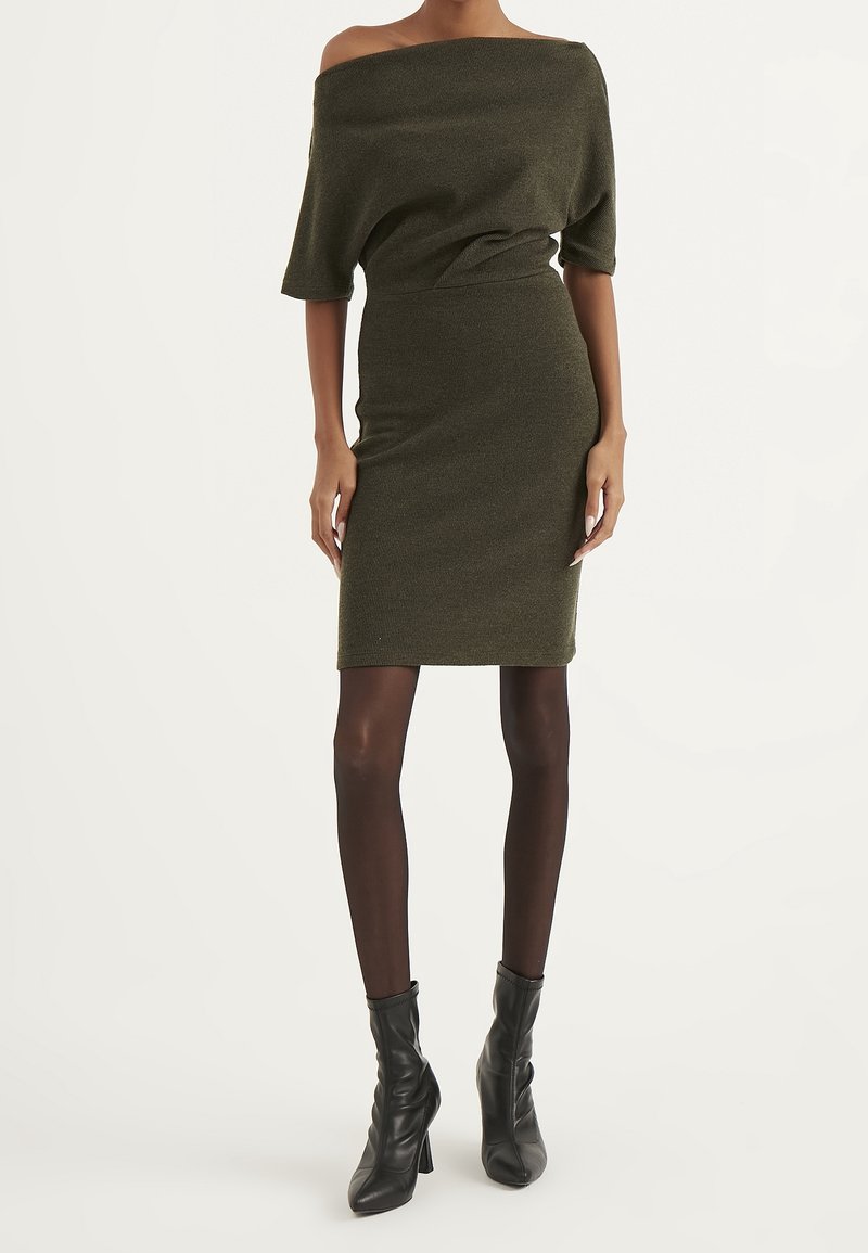 Olivengrünes Strickkleid mit schulterfreien Trägern, taillierter Silhouette und Rüschen-Details, kombiniert mit schwarzen Stiefeletten und transparenten Strumpfhosen.