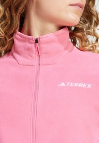 Chaqueta de forro polar con cremallera en rosa intenso, que presenta un cuello alto y un sutil logotipo en blanco. La tela tiene un acabado suave y texturizado.