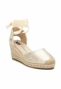 Zapato de cuña espadrille dorado con punta redondeada, detalles de yute tejido y cintas en el tobillo. Presenta un acabado metálico suave y lados recortados.