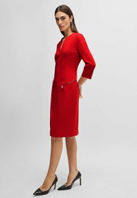 Rotes knielanges Kleid mit V-Ausschnitt, Dreiviertelärmeln und Reißverschlusstaschen. Glattes Material und taillierte Form, kombiniert mit schwarzen Absätzen.