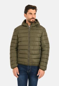 Uomo che indossa una giacca a vento verde oliva con cappuccio e tasche con zip, abbinata a jeans blu scuro, mentre guarda leggermente di lato.