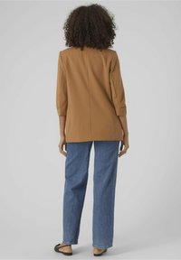 Brązowa, dopasowana marynarka z rękawami do łokcia i rozcięciem z tyłu, zestawiona z wysokim stanem niebieskich dżinsów i czarnych płaskich sandałów.