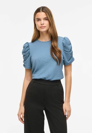 Femme aux longs cheveux bruns portant un haut bleu texturé à manches bouffantes et un pantalon noir taille haute, debout devant un fond uni.