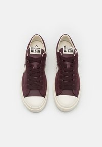 Vinröda canvas låga sneakers med vitt gummitäcke på tån och snören, med en strukturerad yta och "Converse All Star"-logotypen på tungan.