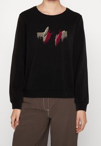 Černá mikina s výšivkou textu ve zlaté, červené a černé barvě s motivem kapky. Dlouhé rukávy a volný střih s žebrovaným lemem.