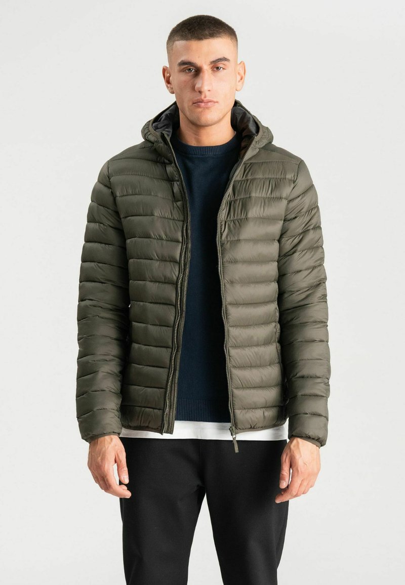 Jeune homme portant une doudoune à capuche vert olive par-dessus un pull bleu marine et une chemise blanche, debout devant un fond clair uni.