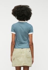 Blå kortærmet top med hvid krave og manchetter, parret med en lysebeige nederdel med to baglommer og et lille logo-patch.